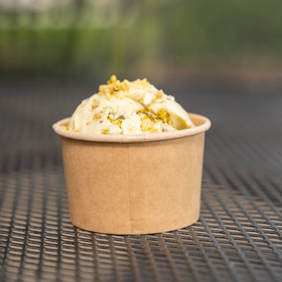 Pistachio ice cream topped with crushed nuts in a brown paper cup on an outdoor metal table.