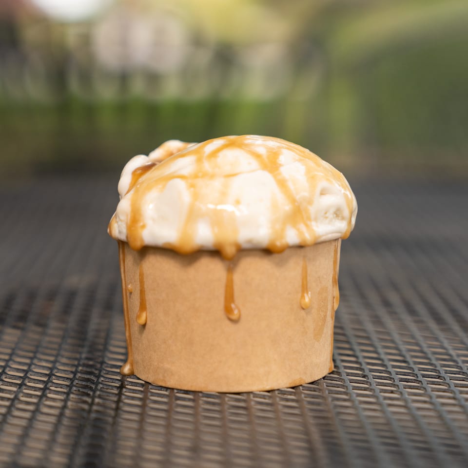 Vanilla ice cream in a cup topped with caramel sauce, resting on a woven outdoor table.